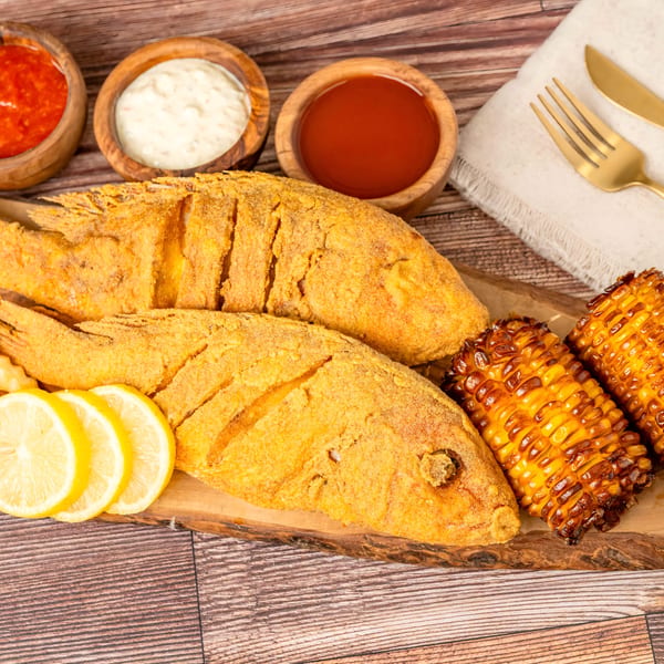 Two whole fried fish with fries, corn on the cob, lemon slices, and dipping sauces on a wooden board.