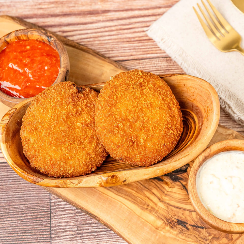 Two golden-brown fried croquettes in a wooden bowl, with red and white dipping sauces on a wooden board.