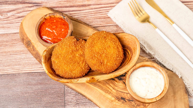 Two golden-brown fried croquettes in a wooden bowl, with red and white dipping sauces on a wooden board.