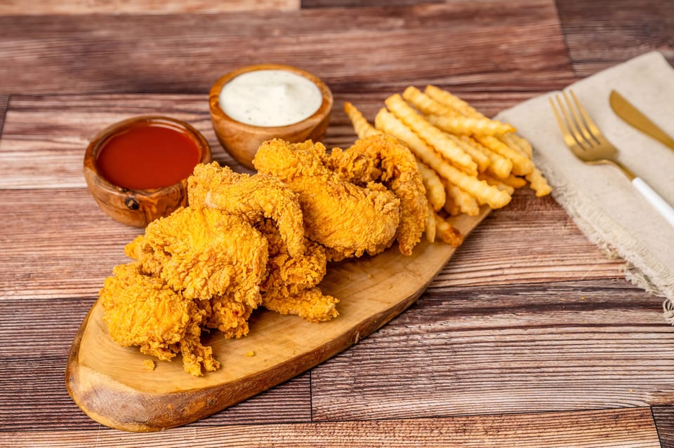Crispy fried chicken tenders and crinkle-cut fries on a wooden board with dipping sauces.