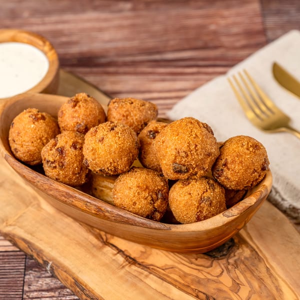 A wooden bowl of golden-brown hushpuppies with a side of white dipping sauce on a wooden board.