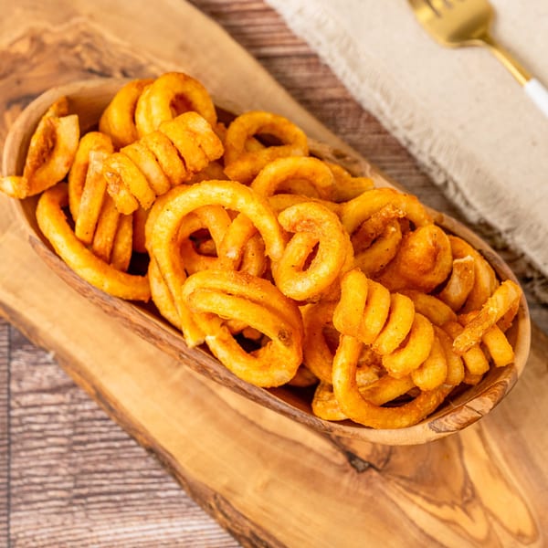 A wooden bowl filled with golden curly fries sits on a rustic wooden board with cutlery.