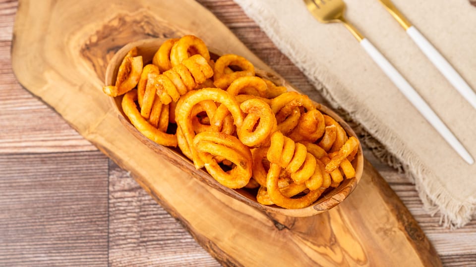 A wooden bowl filled with golden curly fries sits on a rustic wooden board with cutlery.