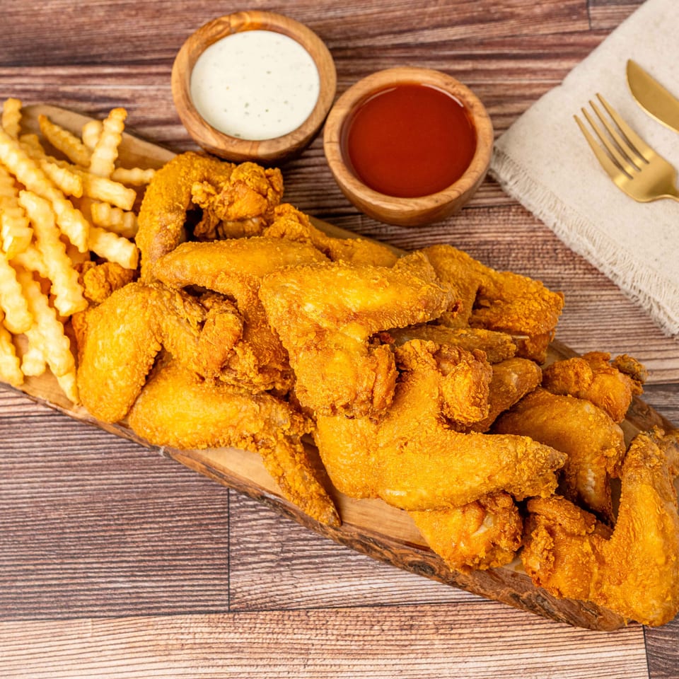 Crispy fried chicken wings and crinkle-cut fries on a wooden board with dipping sauces and cutlery.