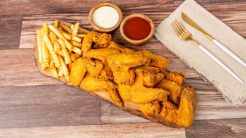 Crispy fried chicken wings and crinkle-cut fries on a wooden board with dipping sauces and cutlery.