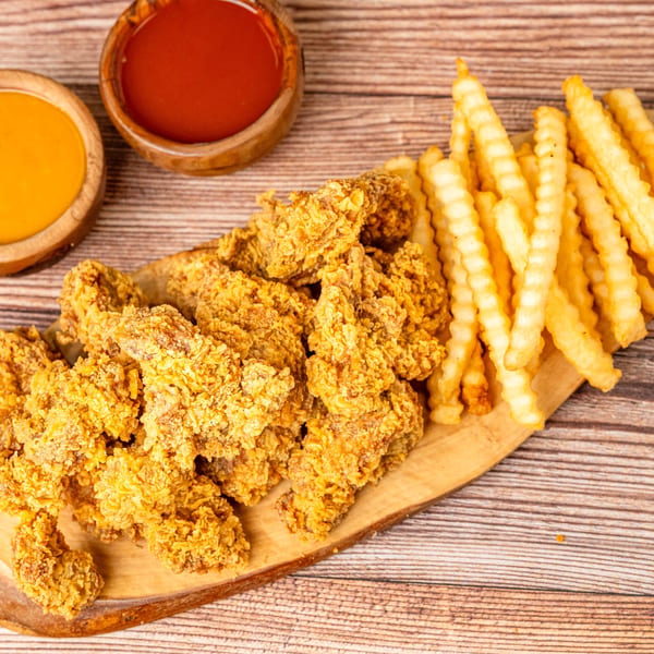Crispy fried chicken tenders and crinkle-cut fries on a wooden board with two dipping sauces.