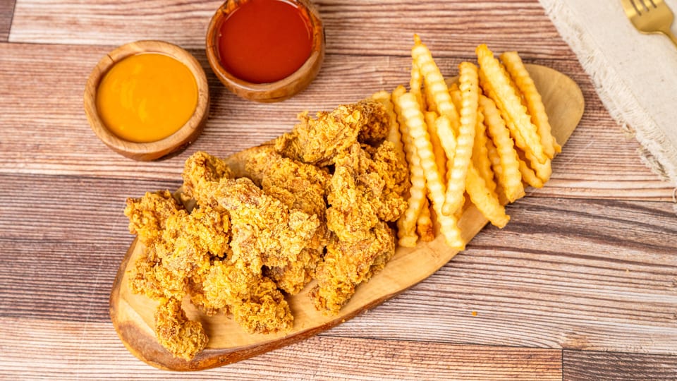 Crispy fried chicken tenders and crinkle-cut fries on a wooden board with two dipping sauces.