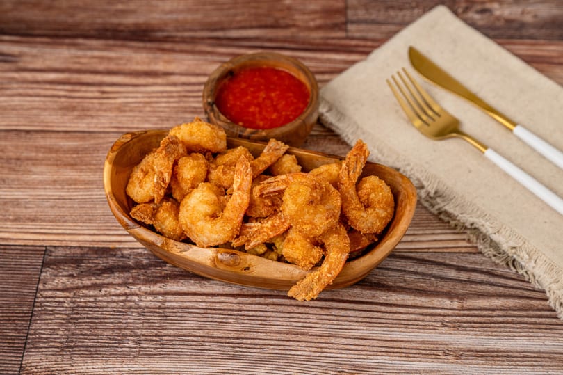 Golden fried shrimp in a wooden bowl, with red dipping sauce and gold cutlery on a napkin.