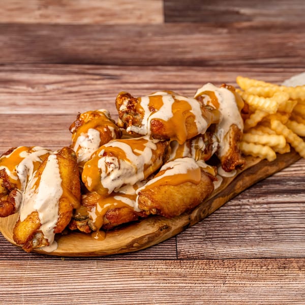 Chicken wings drizzled with orange and white sauces, served with crinkle-cut fries on a wooden board.