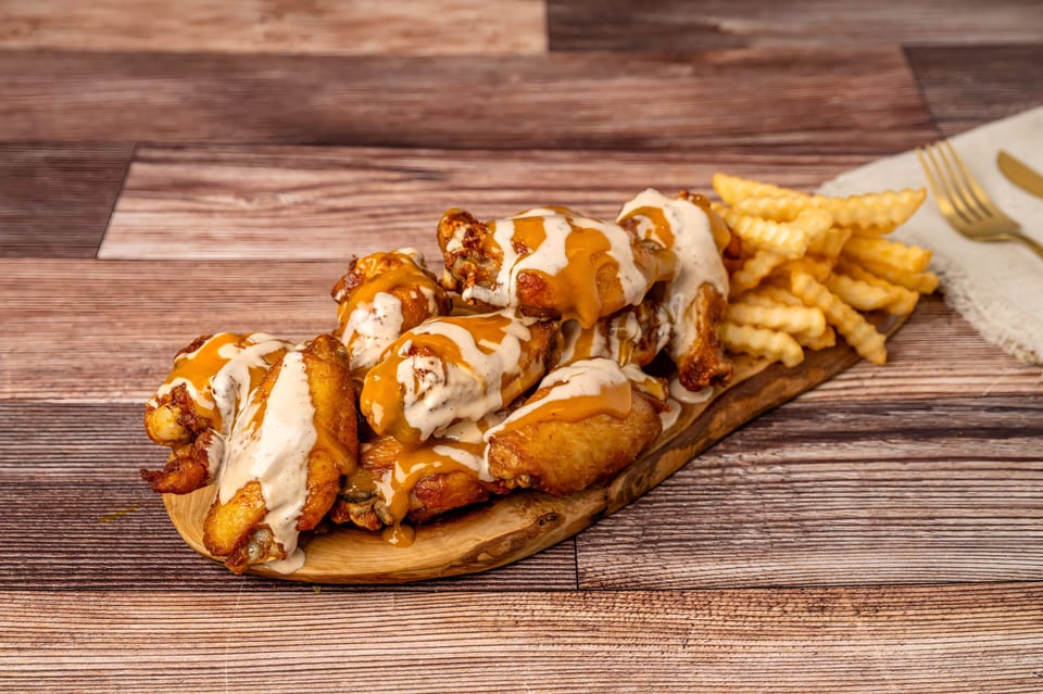 Chicken wings drizzled with orange and white sauces, served with crinkle-cut fries on a wooden board.